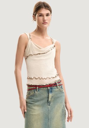 Giovane donna che indossa un top crema con volants e una gonna di jeans blu sbiadita con una sottile cintura rossa, in piedi davanti a uno sfondo neutro.