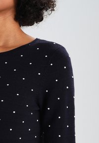 Granatowy sweter z okrągłym dekoltem, ozdobiony białymi groszkami. Wykonany z miękkiego materiału, o dopasowanym kroju i prążkowanej fakturze.