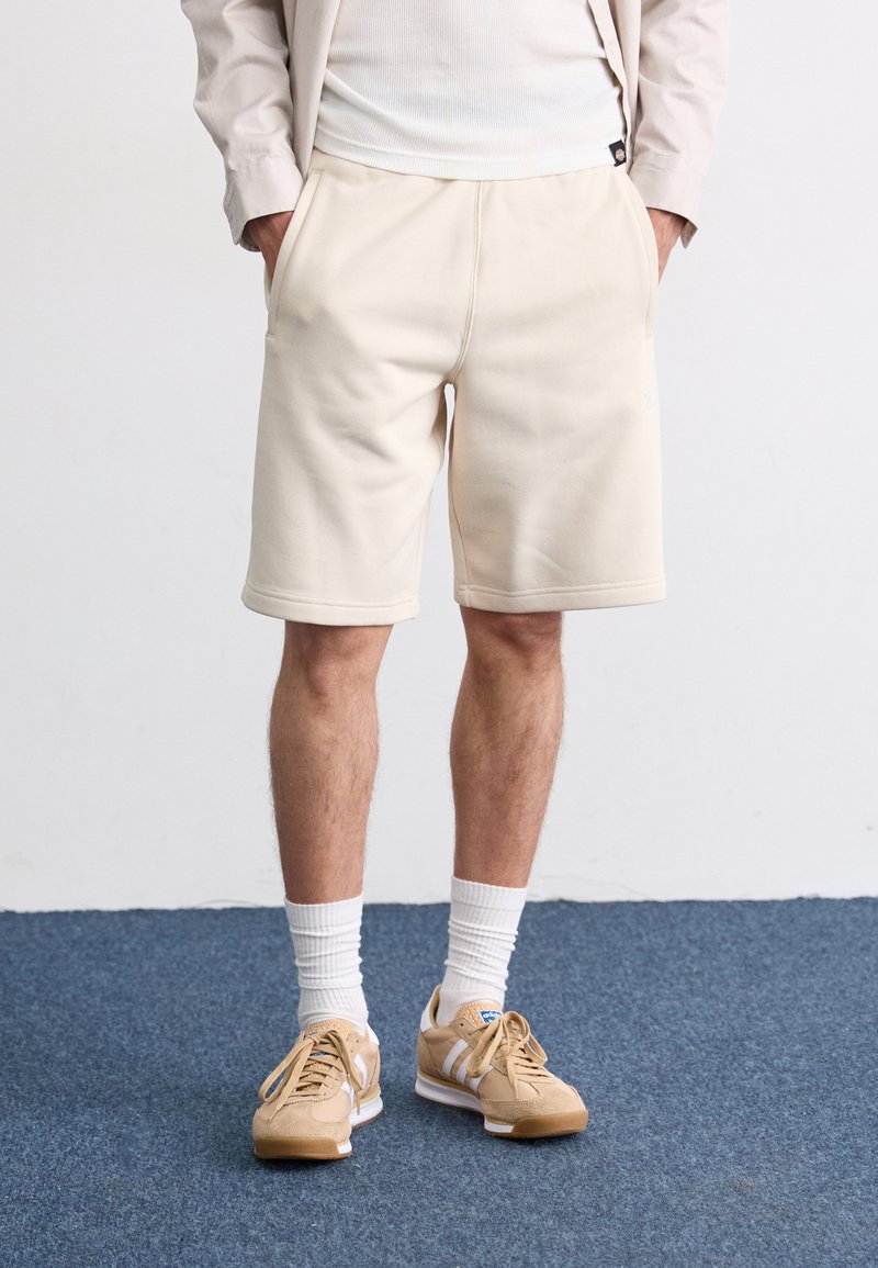 Light beige shorts made of soft fabric with side pockets, featuring a relaxed fit and a flat hem. Paired with tan sneakers and white socks.
