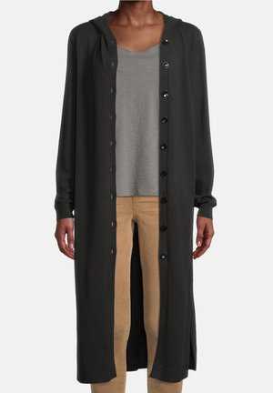 Personne portant un long cardigan noir boutonné avec capuche, un t-shirt gris et un pantalon beige, debout devant un fond uni.