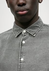 Camisa de ganga cinza, com colarinho, cinco botões brancos e detalhes de costura visíveis, apresentando uma textura suave e um brilho subtil.