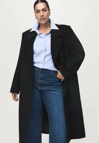 Abrigo negro de corte doble, con solapas anchas, llevado sobre una camisa de rayas azul claro y jeans azules de talle alto. Tejido suave, ajuste estructurado.