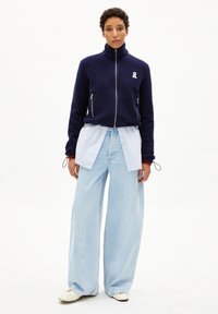 Veste en polaire navy avec un col haut et un logo en ruban blanc, associée à un pantalon en jean large bleu clair et des chaussures crème.