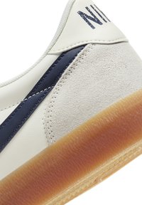 Sapatilhas brancas com detalhes em azul marinho, apresentando um cabedal em couro liso e um painel em camurça texturizada. Sola em borracha de borracha com um padrão de ranhuras.