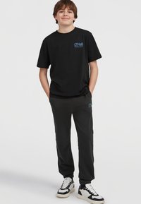 Černé bavlněné tričko s modrým logem na hrudi, spárované s tmavě šedými tepláky. Bílé a černé tenisky doplňují celý outfit.