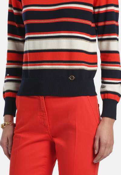 Pull rayé aux couleurs rouge, marine et blanc, avec des poignets côtelés. Assorti à un pantalon rouge vif et un accent doré à la taille.