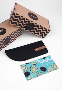 Černý plstěný obal s koženým popruhem, doplněný o dvě vzorované balicí krabice s cik-cak vzorem a jednou krabicí s motivem zeleného kiwi.