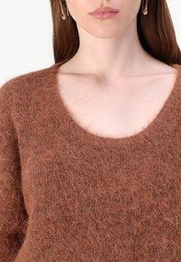 Rjava mohair pulover z mehkobno, puhasto teksturo. Ima širok krojen izrez in dolge rokave. Zlahka oblečljiv in z vročim videzom.