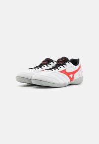 Scarpe sportive bianche con lacci neri, accenti rossi e un design strutturato. Presentano una punta arrotondata e una suola in gomma per una maggiore aderenza.
