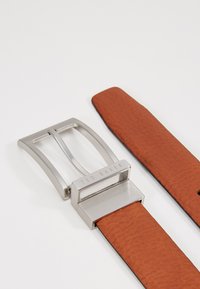 Cinturón de cuero marrón con una superficie texturizada, que cuenta con una hebilla de metal plateado grabada con "TED BAKER". Diseño minimalista con forma rectangular.