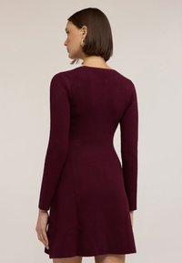 Abito lungo di colore bordeaux con maniche lunghe, caratterizzato da un corpetto aderente, gonna svasata e scollatura rotonda, realizzato in un tessuto liscio. Orecchini dorati visibili.