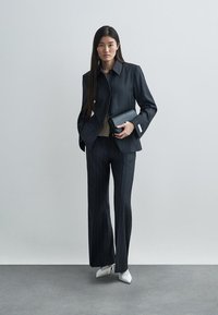 Kohandatud tume triibuline ülikond lehvikpükstega ja kraega jakk. Sobitatud hele tekstureeritud topi ja musta käekotiga. Valged kontsakingad.