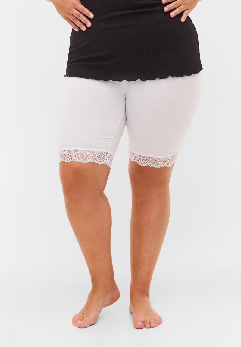 Femme portant un short blanc mi-cuisse avec une bordure en dentelle et un haut noir, debout pieds nus sur un fond uni.