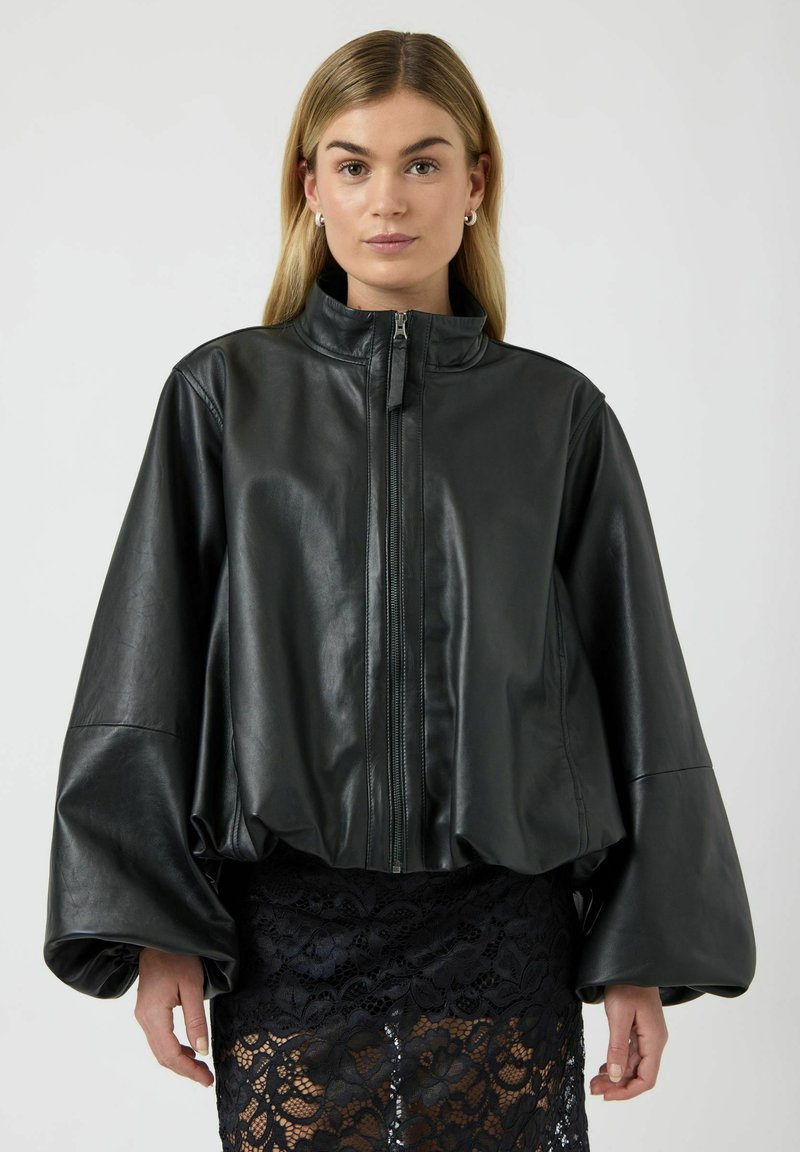 Veste courte en cuir noir avec un col montant et des manches oversize, dotée d'une fermeture éclair à l'avant et d'une texture lisse. Portée sur une jupe en dentelle.