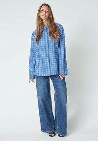 Modra karirana oversized srajca z dolgimi rokavi, v kombinaciji z širokimi modrimi usnjenimi hlačami in črnimi sandalami na peto. Enostavna ozadje.