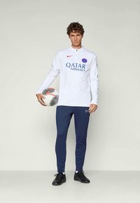 Junger Mann in Paris Saint-Germain Trainingsoberteil und blauen Hosen, der einen Fußball vor einem einfarbigen Hintergrund hält.