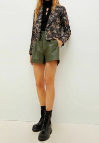 Sorte mønstrede cropped jakke, tætsiddende sort turtleneck, grønne shorts i kunstlæder med frontknapper og sorte snørede kampstøvler.