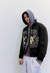 Musta pommitakki, jossa graafinen kuviointi ja harmaa ruudullinen hupullinen osa, kerrospukeutuneena viininpunaisen villapaidan päälle, yhdistetty vaaleanharmaisiin collegehousuihin.