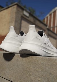 Coppia di scarpe da ginnastica bianche Adidas Ultraboost posate su un bordo di pietra all'aperto con un edificio in mattoni sfocato sullo sfondo.