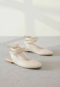 Beige flats with a rounded toe, soft leather material, and a transparent strap design. Slit sides with knotted accent details. Flat sole.