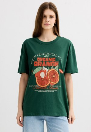 Mujer joven con camiseta de color verde oscuro con gráfico de fruta de naranja y texto "Organic Orange", combinada con vaqueros azul claro, de pie sobre fondo blanco.