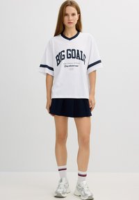 Camiseta blanca de gran tamaño con texto en azul marino "GRANDES OBJETIVOS", de mangas cortas, con cuello en V; combinada con una falda plisada en azul marino y zapatillas blancas.