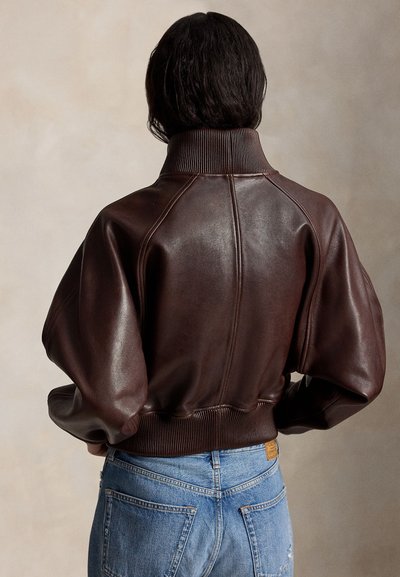 Άτομο με κοντά σκοτεινά μαλλιά που φορά σκούρο καφέ δερμάτινο μπουφάν τύπου bomber και μπλε τζιν, γυρισμένο προς τα πίσω μπροστά σε απλό φόντο.