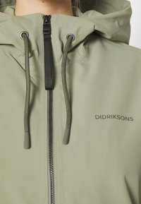 Veste vert clair avec fermeture éclair, capuche à cordon de serrage et logo. Texture lisse avec des accents noirs sur la fermeture éclair et les cordons.