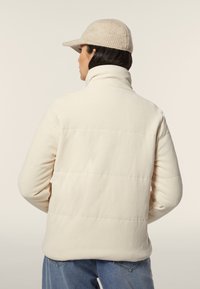 Chaqueta acolchada cremosa con textura de canalé vertical, cuello alto y silueta ajustada. Llevada con un gorro de punto ligero y vaqueros azules.