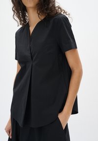 Top corto nero a maniche corte con scollatura a V e dettaglio plissettato sul davanti. Realizzato in materiale leggero, presenta una vestibilità rilassata e un orlo arcuato.