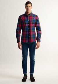 Camisa de cuadro con botones que presenta patrones en rojo, verde y azul. Cuello clásico y mangas largas combinadas con jeans oscuros y zapatos marrones.