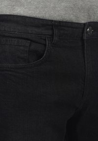 Pantalones vaqueros de mezclilla negra con una textura suave y costuras visibles, que cuentan con un bolsillo y un cierre de botón metálico en la cintura.