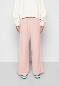 Pantalones de pierna ancha en rosa, de tela suave, que presentan un corte relajado y un detalle de logo. Combinados con una sudadera oversized en crema y zapatillas.