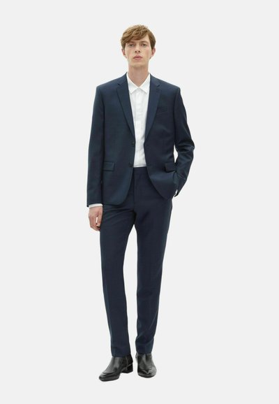 Granatowy garnitur z dopasowaną marynarką i pasującymi spodniami. Pod spodem znajduje się biała koszula oraz czarne botki za kostkę. Minimalistyczny design.