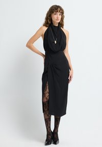 Czarna sukienka na halterze z drapowanym korpusem i bocznym rozcięciem, w połączeniu z czarnymi rajstopami w kwiatowy wzór z koronką i błyszczącymi czarnymi szpilkami.