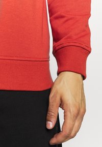 Sweat-shirt rouge avec poignets et taille côtelés, présentant une texture lisse. Main posée près de l'ourlet, mettant en valeur la coupe du vêtement.