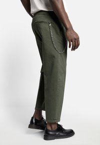 Mand iført olivengrønne bukser med en sølvkæde som tilbehør og sorte læder snørebåndssko, stående på en hvid baggrund.