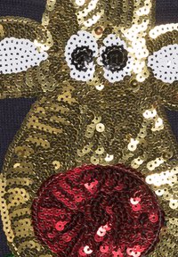 Ornement de sequins dorés et rouges représentant un visage d'animal stylisé sur un fond de tissu sombre, avec des détails en noir et blanc.