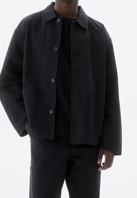 Black canvas jacket with a collared neck, front buttons, and two side pockets. Worn over a black shirt, with a relaxed fit.