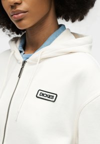 Κοντινό πλάνο ατόμου που φοράει λευκό φούτερ με φερμουάρ και μαύρο λογότυπο "Dickies" και μπλε γιακά πουκάμισου ορατό από κάτω.