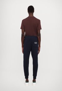 Ruskea puuvillainen t-paita yhdistettynä laivastonsinisiin collegehousuihin, joissa on takatasku ja valkoinen logo. Mallilla on valkoiset lenkkarit vihreillä yksityiskohdilla.