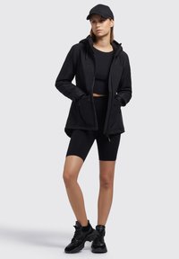 Giacca sportiva nera con cappuccio, chiusura zip e tasche, abbinata a un crop top nero e pantaloni biker coordinati, completata da sneakers nere.