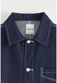 Veste en denim foncé avec des coutures blanches, un col classique, une fermeture à boutons-pression à l'avant et une poche poitrine unique. L'étiquette affiche le nom de la marque.