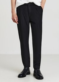 Pantaloni neri su misura con pieghe, realizzati in tessuto liscio. Presentano una vestibilità slim e una fibbia lucida. Abbinati a scarpe di pelle nere.