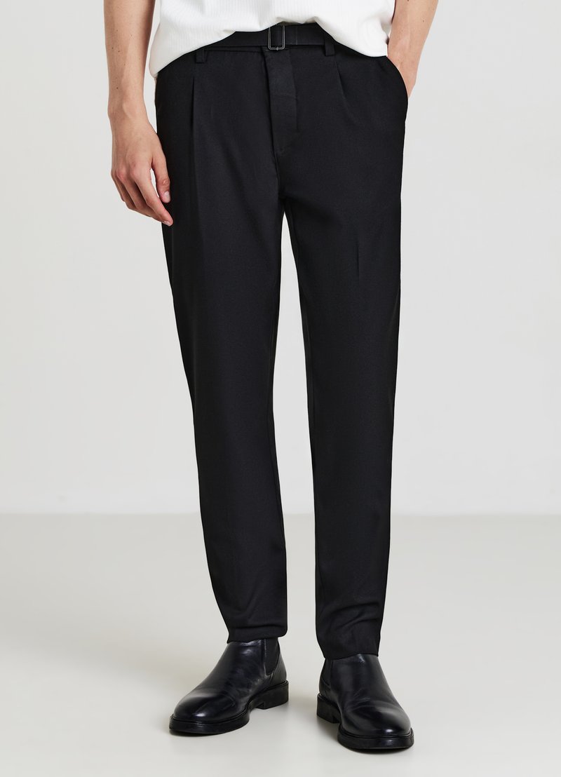 Pantaloni neri su misura con pieghe, realizzati in tessuto liscio. Presentano una vestibilità slim e una fibbia lucida. Abbinati a scarpe di pelle nere.