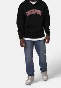 Sudadera negra con texto rojo "CRYSTALPARIS", con bolsillo delantero, llevada sobre una camiseta blanca, combinada con jeans azules y zapatillas grises.