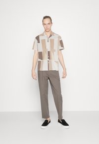 Camisa de manga corta con patrones geométricos abstractos en beige y marrón, con botones en la parte frontal, combinada con pantalones grises y zapatos negros sin cordones.