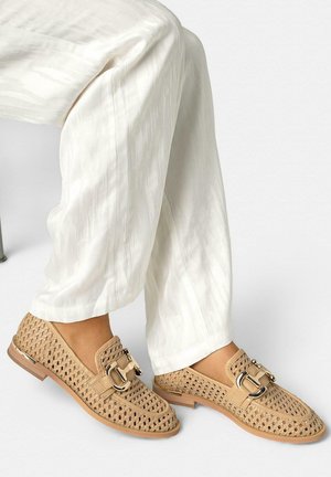 Beige geweven loafers met gouden gespdetail, gedragen met witte textuurwijde broekspijpen bij een zittend persoon.