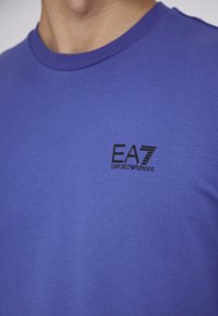 T-shirt di cotone viola con colletto a girocollo. Presenta un logo EA7 Emporio Armani nero sul petto. Texture liscia, design aderente.