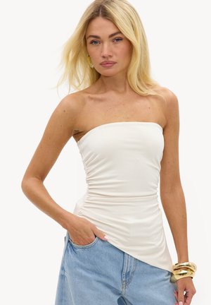 Femme blonde portant un haut blanc froncé sans bretelles et un jean bleu clair, accessoirisée de bracelets et boucles d'oreilles dorés, posant avec la main dans la poche.